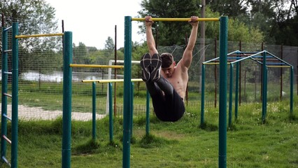 Athletic man trains abdominal muscles raising legs on horizontal bar at sports ground. Young sportsman exercising on pull-up bar outdoor. Sporty athlete working out at nature. Healthy lifestyle
