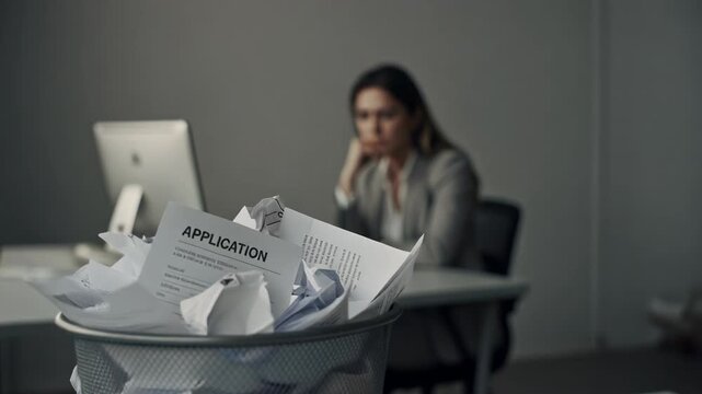 Frustrated woman in office with rejected job applications in trash bin depicting job search stress and career challenges