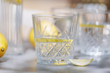 Refreshing summer drink in a crystal glass with lemon slice, whole lemons and pitcher in the background on an outdoor gray table