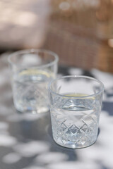 Two transparent glasses filled with water and lemon slices, placed on a gray patio table with leafy tree shadows and warm natural light