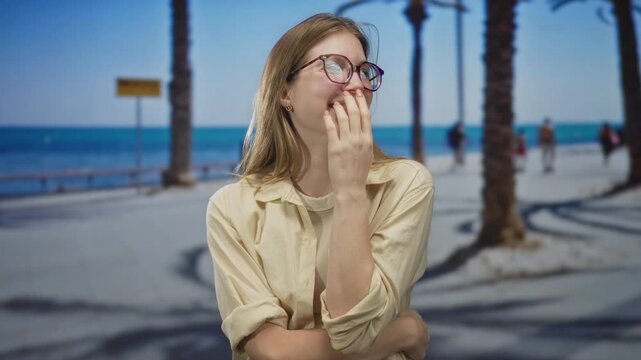 Young blonde woman covers mouth while laughing amid palm trees on a sunlit seaside promenade; carefree joy.