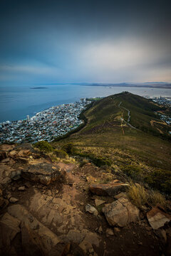 Aerial view of a sprawling city meeting the ocean under a dramatic sky, framed by rugged mountain terrain and rocky outcrops, Cape Town, Western Cape, South Africa.