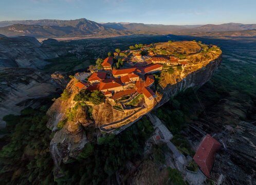 Aerial view of red-roofed Meteora monastery complex perched atop towering rock formations, bathed in the warm glow of the setting sun, Kalabaka, Greece.