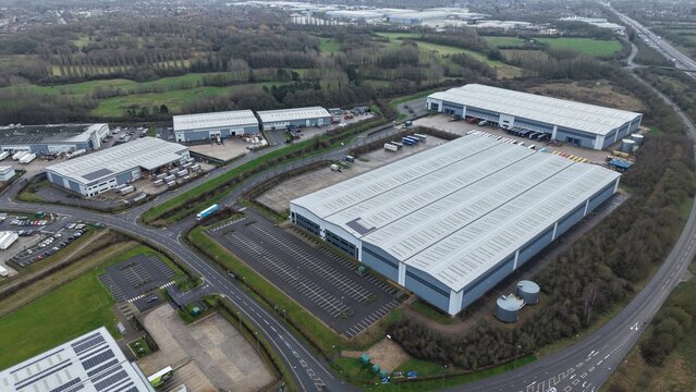 Glenfield, United Kingdom - 08 February 2026: Aerial view of industrial warehouses casting long shadows under a grey sky, surrounded by patches of green fields and winding roads.