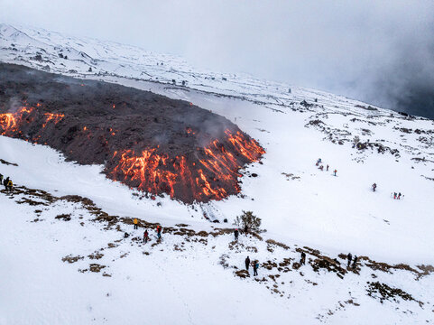 Aerial view of fiery lava flows down the snow-covered slopes of Mount Etna, contrasting the stark white landscape with intense orange hues, Catania, Sicily, Italy.