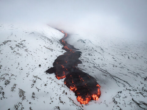 Aerial view of molten lava snakes its way through a stark white, snow-covered landscape, a fiery river cutting through the frozen stillness, Catania, Sicily, Italy.