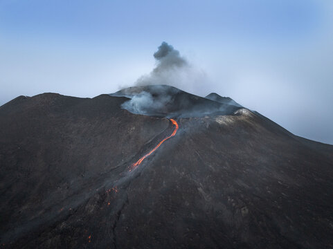 Aerial view of fiery lava flows down the dark, volcanic slopes under a hazy sky, a stark contrast of heat and ash, Mount Etna, Catania, Sicily, Italy.