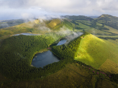 Aerial view of twin lakes nestled amidst verdant hills, where sunlight kisses the slopes and mist veils the peaks, creating an ethereal landscape, Miradouro do Pico do Carvao, Acores, Portugal.