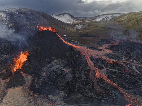 Aerial view of fiery lava flows down rugged, dark slopes, contrasting sharply with the surrounding snow-dusted green hills under a stormy sky, Grindavik, Iceland.