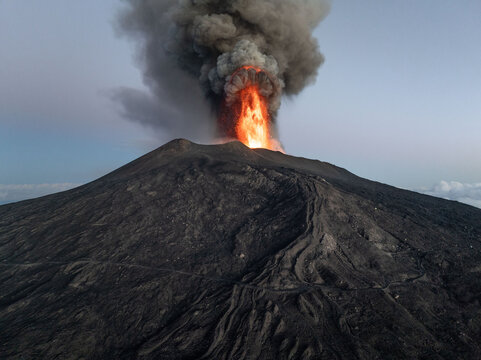 Aerial view of fiery lava erupts from the summit of Mount Etna, contrasting against the dark volcanic rock and the soft, dusky sky, Catania, Sicily, Italy.