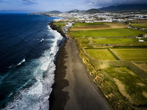 Aerial view of the stark contrast between the black sand beach, the deep blue ocean, and the vibrant green fields stretching towards the town, Santa Barbara, Azores, Portugal.