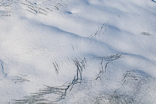 Aerial view of a stark, white expanse crisscrossed with dark, jagged fractures, revealing the raw power of the frozen landscape, Greenland.