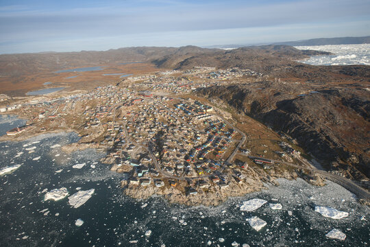 Aerial view of a quaint, colorful town nestled between rugged, rocky hills and an icy fjord dotted with shimmering icebergs, Ilulissat, Greenland.