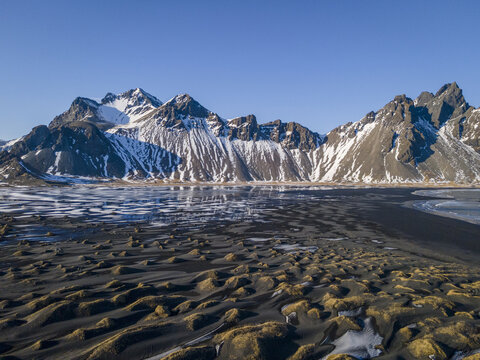 Aerial view of black sand beach contrasting with snow-capped mountains under a clear blue sky, creating a stark but serene landscape, Sveitarfelagid Hornafjordur, Iceland.