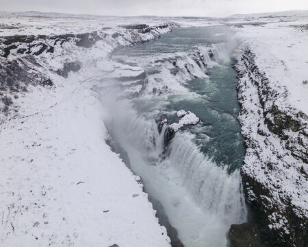 Aerial view of icy waters plunge dramatically over a rocky cliff into a mist-shrouded abyss, framed by snow-dusted landscapes, Hrunamannahreppur, Iceland.