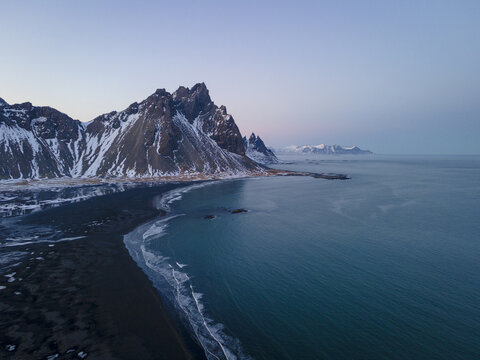Aerial view of Stokksnes' black sand beach kissed by the turquoise sea, cradled by snow-dusted mountains under a pastel sky, Sveitarfelagid Hornafjordur, Iceland.