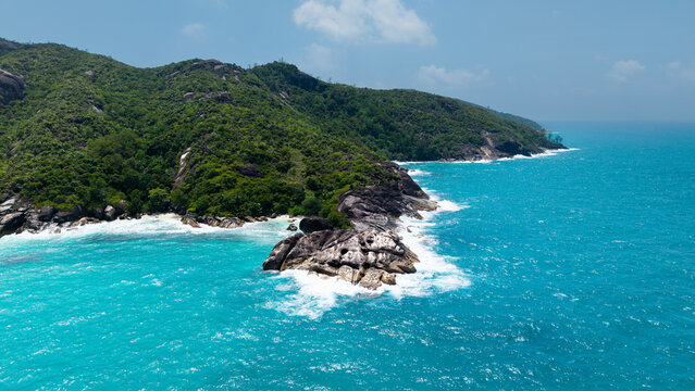 Aerial view of the vibrant turquoise sea colliding with dark, rocky outcrops and lush green hills along Anse Major Trail, Anse Major, Mahe, Seychelles.