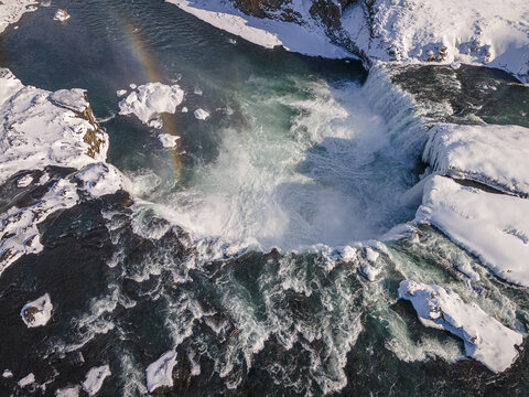 Aerial view of the powerful waterfall cascading into the river below, surrounded by snow-covered rocks and a faint rainbow, Pingeyjarsveit, Iceland.