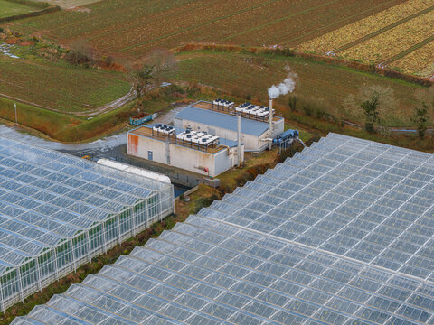 Aerial view of a modern facility nestled amidst a grid of greenhouses, smoke gently rising against a backdrop of patterned fields, Tredarzec, Bretagne, France.
