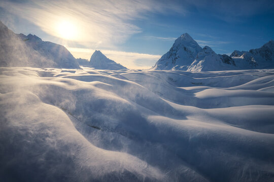 Aerial view of a windswept snowscape, where icy gusts sculpt the drifts into fluid forms beneath the watchful gaze of snow-capped peaks, Valdez, Alaska, United States.