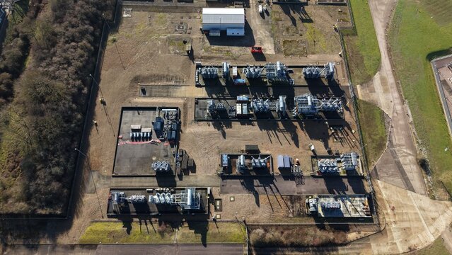 Aerial view of an industrial zone with metallic structures casting stark shadows on the gravel ground, framed by patches of green grass and dense trees, Luton, United Kingdom.