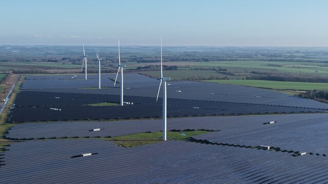 Aerial view of wind turbines standing tall amidst dark solar panels contrasting against the verdant fields stretching to the horizon, Chelverston, Wellingborough, United Kingdom.