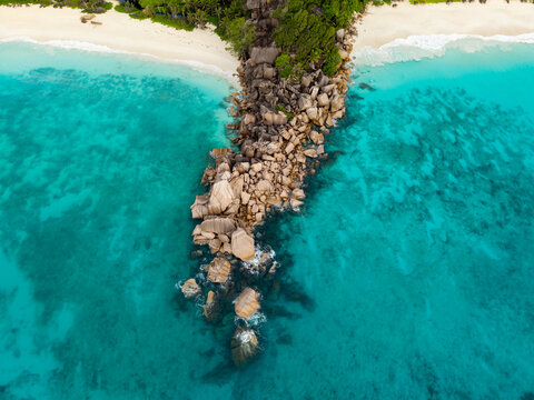 Aerial view of azure waters embracing rugged rocks and lush foliage along the coast, creating a striking contrast of textures and tones, Anse Reunion, La Digue, Seychelles.