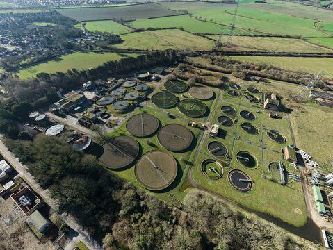 Aerial view of circular sewage tanks stand in stark contrast to the surrounding green fields and bare trees at the sewage works, Luton, United Kingdom.