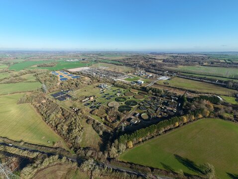 Aerial view of circular sewage tanks contrasting with the surrounding green fields and distant treeline under a clear blue sky, Luton, Luton, United Kingdom.