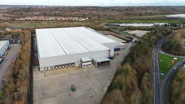 Northampton, United Kingdom - 14 February 2026: Aerial view of a vast, modern distribution warehouse with a gleaming white roof contrasting against the surrounding muted green and brown landscape.