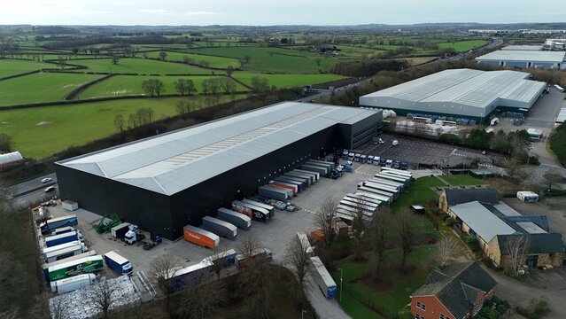 Northampton, United Kingdom - 14 February 2026: Aerial view of the vast, dark-toned warehouse complex contrasting with the bright, verdant fields beyond, a testament to modern logistics.