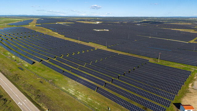 Aerial view of solar panels stretching across the landscape like dark mirrors reflecting the sky, bisected by roads and bordered by vibrant green fields, Caceres, Caceres, Espana.