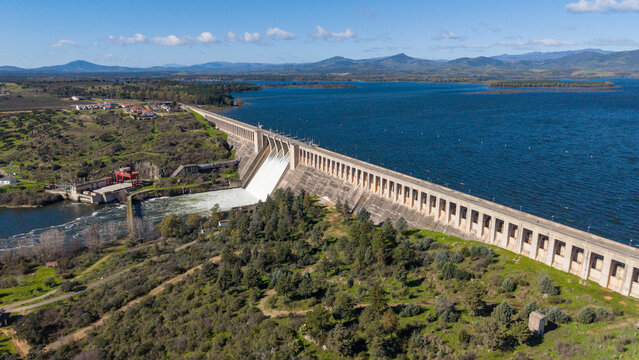 Aerial view of the dam holding back the Embalse de Alcantara, the water reflecting the sky like a mirror, with the green landscape contrasting against the blue, Caceres, Caceres, Espana.