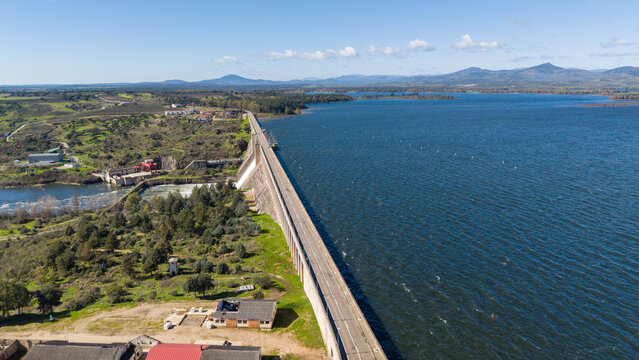 Aerial view of the Embalse de Valdecanas, a vast reservoir reflecting the clear sky, contrasted by the rugged dam and lush greenery, Caceres, Caceres, Espana.