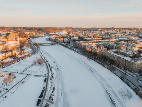 Aerial view of the snow-covered Neris River snaking through the heart of the city, framed by the Gediminas' Tower and the cityscape bathed in the soft glow of winter sunrise, Vilnius, Lithuania.