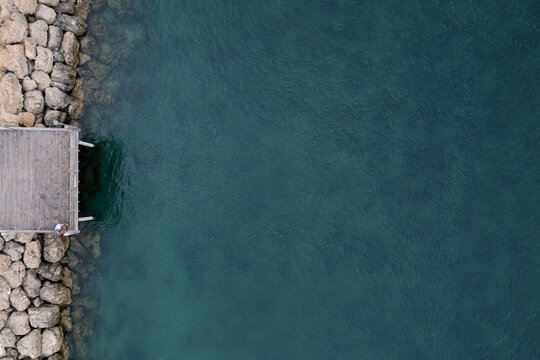 Aerial view of a weathered wooden pier jutting into the teal-blue water, bordered by rugged, sun-kissed rocks, Mandurah, Western Australia, Australia.