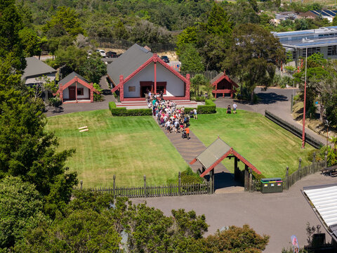 Aerial view of a gathering of people ascending the verdant hill towards the traditional Maori building, its red trim contrasting against the lush greenery, Rotorua, New Zealand.