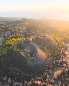 Aerial view of the volcanic cone with a dark, textured surface encircled by vibrant green and gold trees under the warm glow of the sun, Piedimonte Etneo, Sicily, Italy.