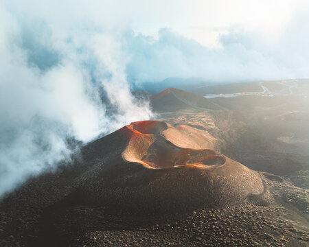Aerial view of a volcanic crater smoldering with fiery intensity against the stark, dark landscape, shrouded in ethereal white clouds, Nicolosi, Sicily, Italy.