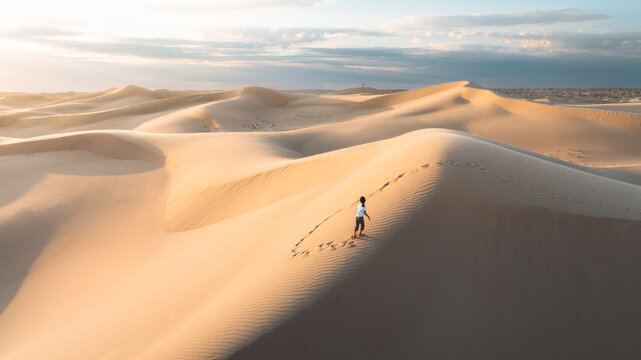 Aerial view of solitary figure traversing undulating dunes under a soft, diffused light, footprints etched in the golden sand, Senek, Mangystau Region, Kazakhstan.