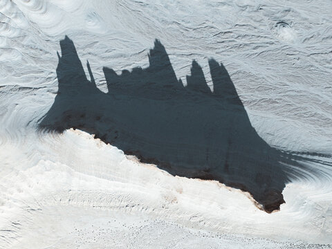 Aerial view of a dark, jagged formation casts long shadows across the stark, rippled landscape, creating a dramatic contrast of light and shade, Aktau, Mangystau Region, Kazakhstan.