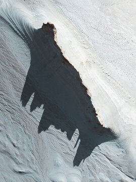 Aerial view of a dark, jagged landform cuts starkly through the pale, textured landscape, creating a dramatic contrast of shadow and light, Aktau, Mangystau Region, Kazakhstan.