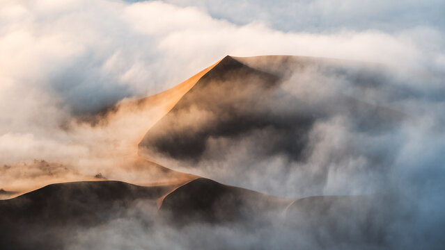 Aerial view of golden dunes piercing through a sea of ethereal fog, a dance of light and shadow in the tranquil desert landscape, Sossusvlei, Hardap Region, Namibia.