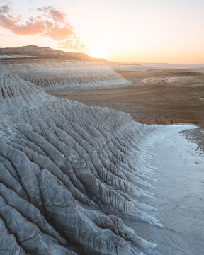 Aerial view of stark, layered rock formations under a soft, peach-tinged sky, painting a serene yet dramatic landscape, Mangystau, Mangystau Region, Kazakhstan.