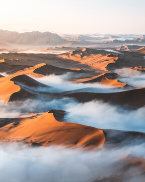 Aerial view of undulating dunes capped with soft, ethereal fog, a dance of warm light and cool mist, Sossusvlei, Hardap Region, Namibia.