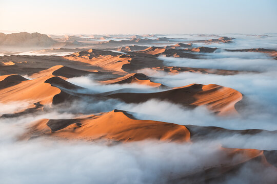 Aerial view of surreal dunes ablaze with sunlight piercing through ethereal fog, creating a dreamscape, Sossusvlei, Hardap Region, Namibia.