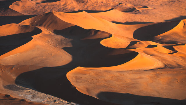 Aerial view of undulating dunes cast stark shadows, their fiery orange hues contrasting sharply against the cool, dark valleys, Sossusvlei, Hardap Region, Namibia.