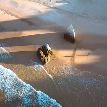 Aerial view of waves gently caressing the sandy shore, framed by striking rock formations casting long shadows in the warm light, Alvor, Faro, Portugal.