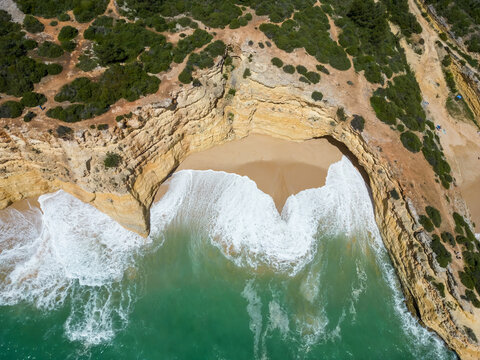 Aerial view of foamy waves crashing on the golden sands nestled below rugged cliffs topped with verdant trees, Praia da Morena, Portugal.