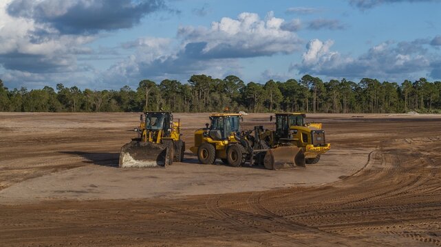 West Palm Beach, United States - 08 November 2024: Aerial view of yellow construction vehicles stand stark against the vast expanse of earth, beneath a sky brushed with wisps of clouds.
